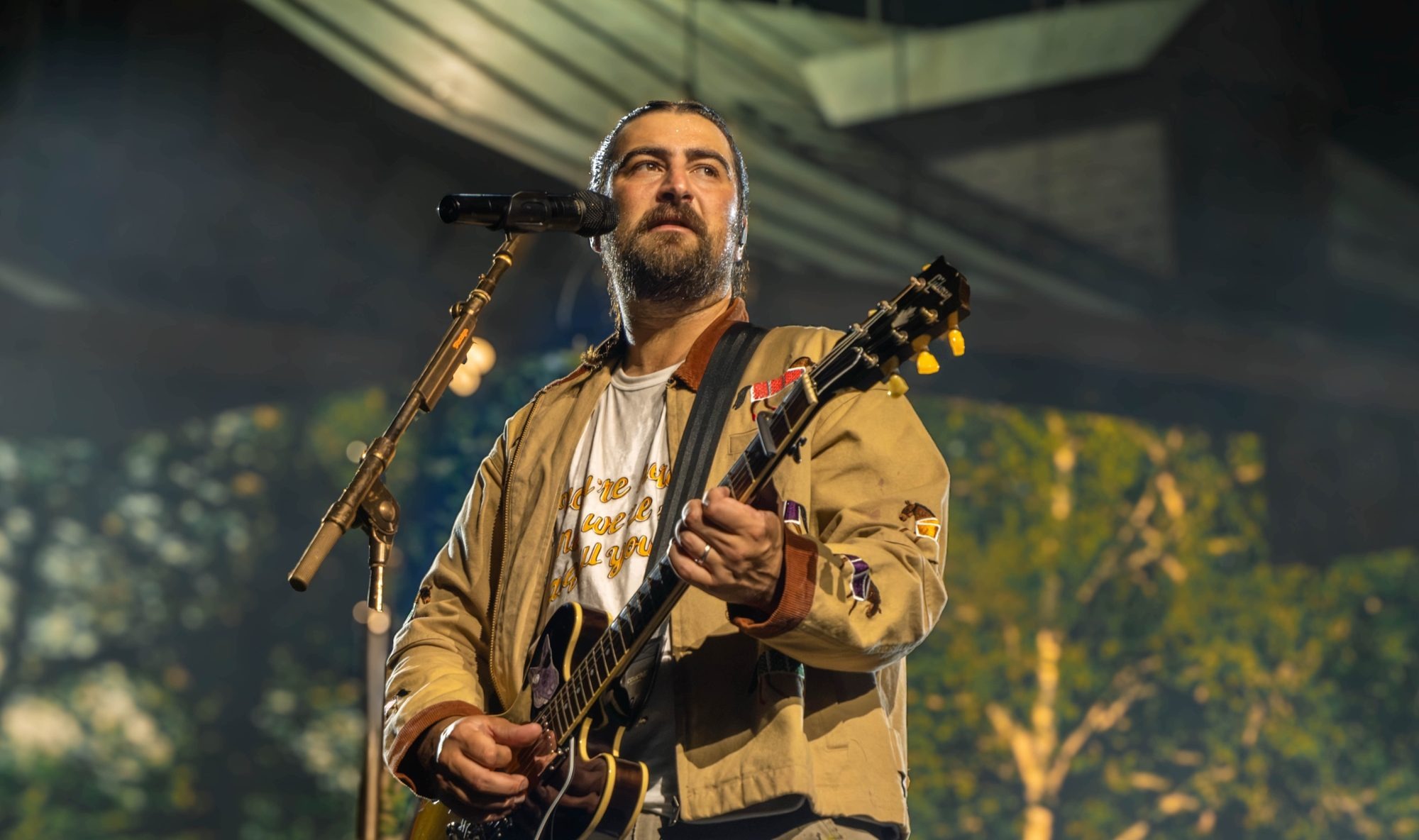 Noah Kahan at the mic with an electric guitar, tree backdrop glowing behind him