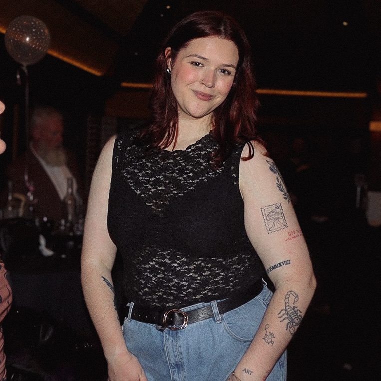 Aimee smiling in a black lace top and jeans, warm lights behind